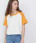 White short-sleeve baseball tee with mustard yellow raglan sleeves and crew neck, worn with blue jeans.