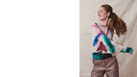 Woman wears a long sleeve silk top in watercolor print with belted metallic trousers