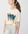 Cream graphic t-shirt with colorful "Hey" text paired with silver pants and a white belt with a large buckle.