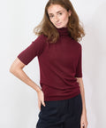Woman wearing a burgundy turtleneck sweater and dark pants against a white background