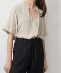 Beige relaxed short-sleeve top with V-neck and subtle pleats, styled tucked into black trousers.