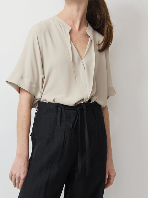 Beige relaxed short-sleeve top with V-neck and subtle pleats, styled tucked into black trousers.