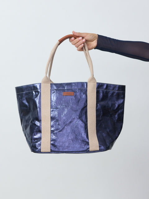 Blue metallic tote bag with beige handles held by a person against a light gray background