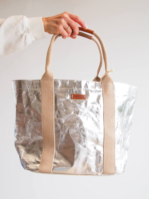 Silver tote bag with beige handles held by a hand on a light background