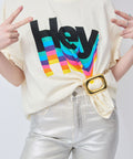 Cream short-sleeve t-shirt with colorful "Hey" graphic, paired with metallic silver pants and a gold belt buckle.