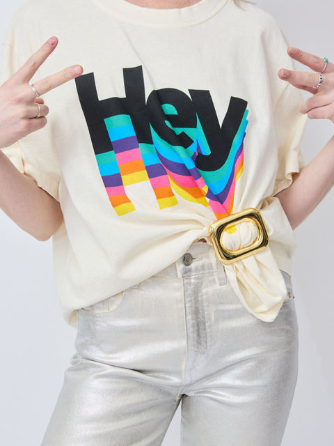 Cream short-sleeve t-shirt with colorful "Hey" graphic, paired with metallic silver pants and a gold belt buckle.