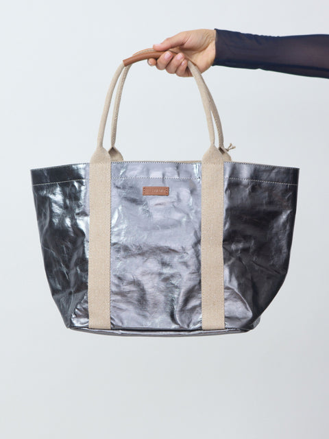 Person holding a metallic silver tote bag with beige handles against a light gray background