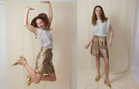 Woman jumping in the air wears a gold pleated bermuda shorts with a white muscle top, gold belt and gold shoes.