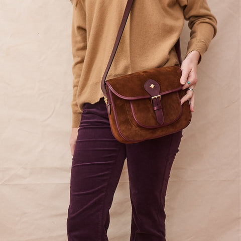 Woman carries a brown suede crossbody bag with leather contrasting trim. Burgundy suede pants and a light brown sweater