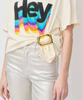 Cream t-shirt with multicolor "Hey" print, tucked into metallic gold pants with a large gold rectangular belt buckle.