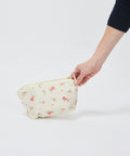 Small ecru makeup pouch with a floral pattern and gold zipper held by hand against a plain background.