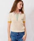 Woman wearing a light-colored knit sweater with yellow accents and blue jeans on a plain background