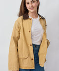 Woman wearing a beige jacket over a white shirt and blue jeans on a plain background