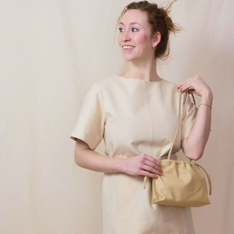 Tall blond wears an 0ff-white mini shift dress with a matching belt. She is holding a cream leather drawstring bag 