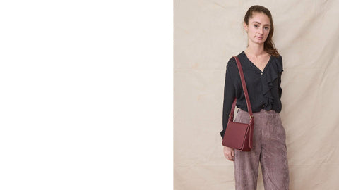 Young woman wears a black blouse with front ruffles, corduroy trousers and a burgundy crossbody leather bag